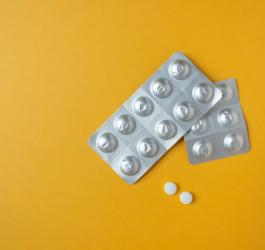 Białe tabletki i srebrne blistry leków na żółtym tle.