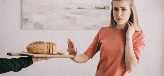 Dziewczyna z alergią pokarmową odsuwa od siebie deskę, na której leży pokrojony chleb.