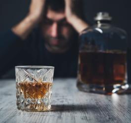 Mężczyzna dotknięty alkoholizmem siedzi przy stoliku, na którym stoi butelka i szklanka z alkoholem.