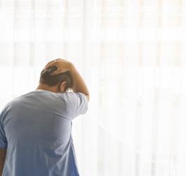 Mężczyzna stoi przy oknie, trzyma się za tył głowy.