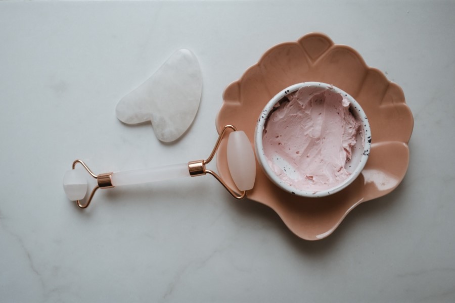 Roller i kamień gua sha do masażu twarzy oraz miseczka wypełniona kremem lub maseczką do twarzy.