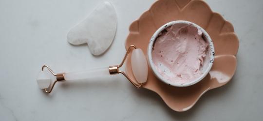 Roller i kamień gua sha do masażu twarzy oraz miseczka wypełniona kremem lub maseczką do twarzy.