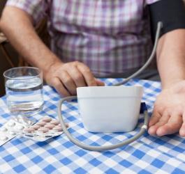 Mężczyzna wykonuje pomiar ciśnienia ciśnieniomierzem. Na stole leżą listy leków i szklanka wody.