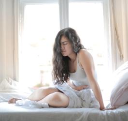 Kobieta siedzi na łóżku, trzyma się za podbrzusze, bo dokuczają jej bóle menstruacyjne.