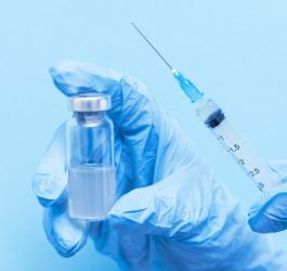 Osoba ma rękawiczki na dłoniach, trzyma w nich fiolkę ze szczepionką oraz strzykawkę.