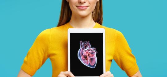 Młoda kobieta trzyma tablet na wysokości klatki piersiowej. Na ekranie wyświetla się model serca.