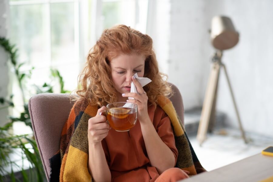 Przeziębiona kobieta siedzi pod kocem, pije herbatę z cytryną, zmaga się z katarem.