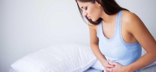 Kobieta siedzi na brzegu łóżka, boli ją brzuch.