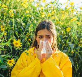 Kobieta w żółtej kurtce, pośród kwiatów. Wydmuchuje nos, bo cierpi na katar alergiczny.