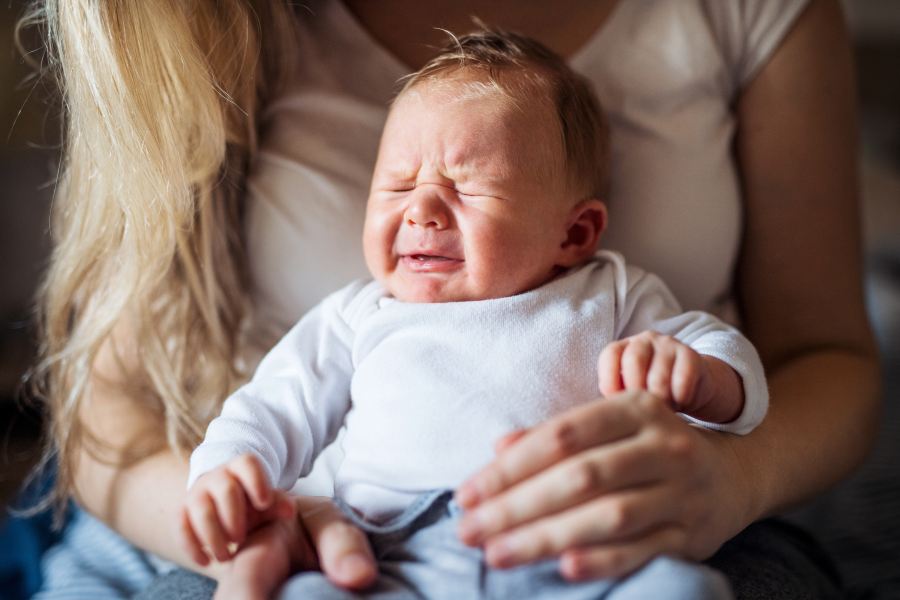 Mama trzyma niemowlę na kolanach. Dziecko ma kolkę niemowlęcą, płacze, jest niespokojne.