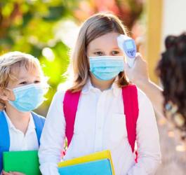 Mali uczniowie w maseczkach. Nauczycielka sprawdza dzieciom temperaturę termometrem bezdotykowym.