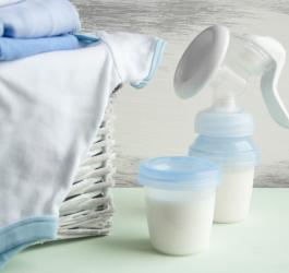 Laktator ręczny i pojemniczek wypełniony odciągniętym mlekiem. Obok kosz z ubrankami dziecięcymi.