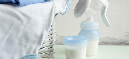 Laktator ręczny i pojemniczek wypełniony odciągniętym mlekiem. Obok kosz z ubrankami dziecięcymi.