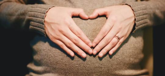 Kobieta trzyma dłonie na ciążowym brzuchu, układa je w kształt serca.