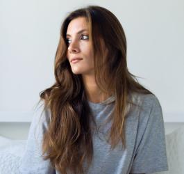 Długowłosa brunetka siedzi na kanapie, spogląda w stronę okna.