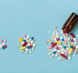 Niebieskie tło. Buteleczka z brązowego szkła, obok rozsypane kolorowe tabletki i kapsułki.