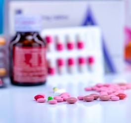 Rozsypane, kolorowe tabletki na blacie. W tle opakowania leków - pudełka, blistry, buteleczki.