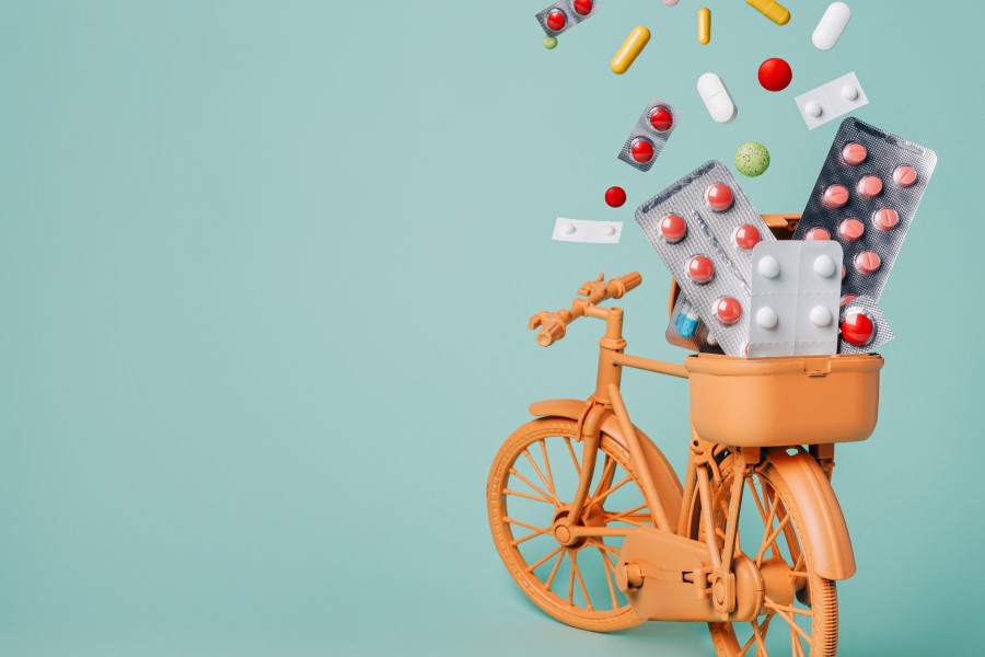 Pomarańczowy, zabawkowy rower przewozi rozmaite leki w tylnym koszyku.