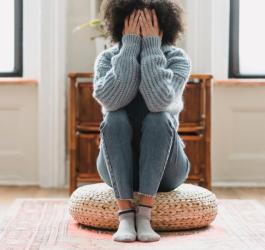 Kobieta siedzi na jutowej pufie, zasłania twarz dłońmi.