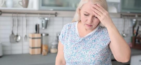 Kobieta w kuchni, trzyma się za głowę, jest obolała i przemęczona.