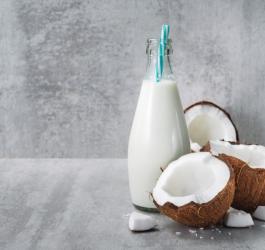 Mleko kokosowe w szklanej butelce oraz rozłupany orzech kokosowy na szarym tle.