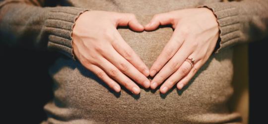 Ciężarna trzyma dłonie na ciążowym brzuchu, palce ułożone są w kształt serca.