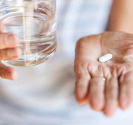 Kobieta trzyma na dłoni tabletkę naproksenu, w drugiej trzyma szklankę wody.
