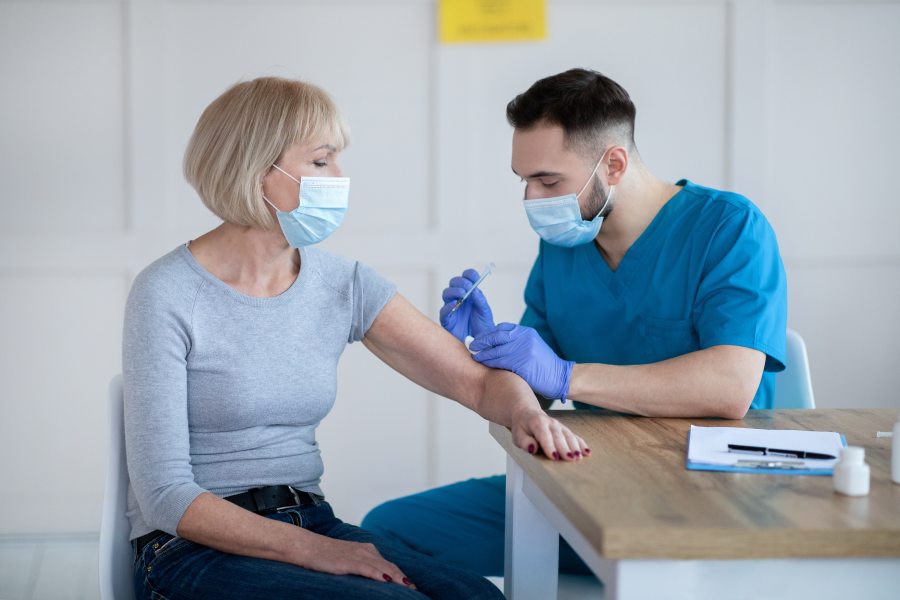 Kobieta zostaje zaszczepiona przeciwko COVID-19 przez pielęgniarza. Oboje są w maseczkach.