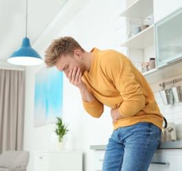 Mężczyzna stoi w kuchni. Trzyma się za brzuch i buzię, odbija mu się po jedzeniu.