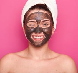 Kobieta z ręcznikiem na głowie i oczyszczającą maseczką na twarzy, uśmiecha się do obiektywu.