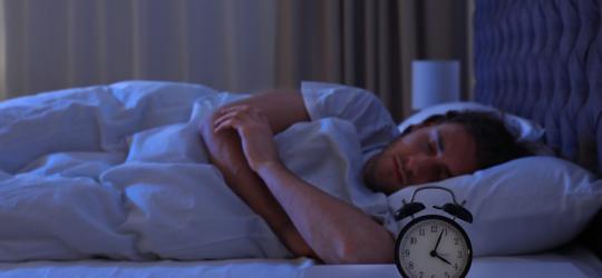 Mężczyzna śpi w łóżku, na stoliku nocnym na ustawiony budzik.