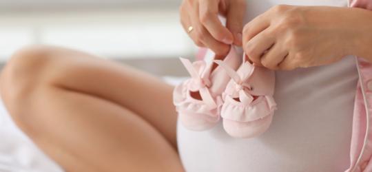 Przyszła mama trzyma na ciążowym brzuchu różowe, niemowlęce buciki.