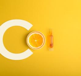 Żółte tło. Litera C, przepołowiona pomarańcza i fiolka serum z witaminą C.