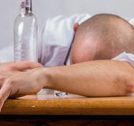 Mężczyzna pod wypływem alkoholu, opiera głowę o blat stołu. Ręką trzyma butelkę z alkoholem.