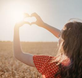 Kobieta układa dłonie w kształt serca na tle zachodzącego słońca.
