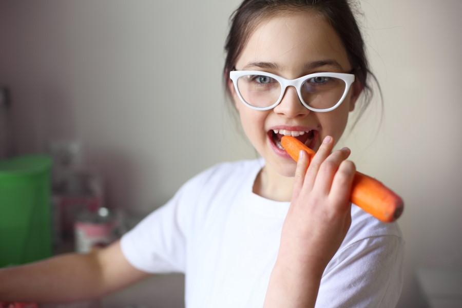 Dziewczynka w okularach zajada się surową marchewką.