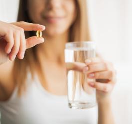Kobieta trzyma w dłoni kapsułkę suplementu, w drugiej ma szklankę wody.