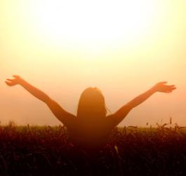 Kobieta stoi na polu pszenicy, na tle zachodzącego słońca, wyciąga ręce ku górze.