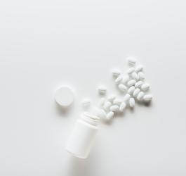 Biała butelka i wysypane z niej białe, owalne tabletki na białym tle.