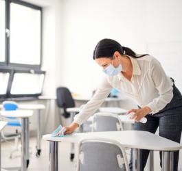 Nauczycielka w maseczce dezynfekuje ławkę w klasie przed rozpoczęciem lekcji.