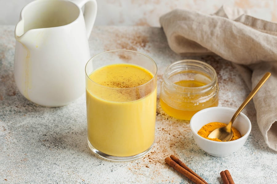 Szklanka złotego mleka obok sproszkowanej kurkumy, miodu i lasek cynamonu.