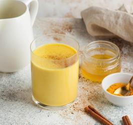 Szklanka złotego mleka obok sproszkowanej kurkumy, miodu i lasek cynamonu.
