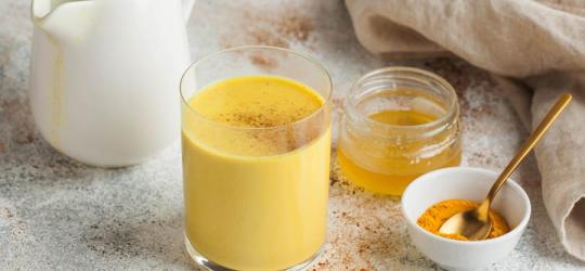 Szklanka złotego mleka obok sproszkowanej kurkumy, miodu i lasek cynamonu.
